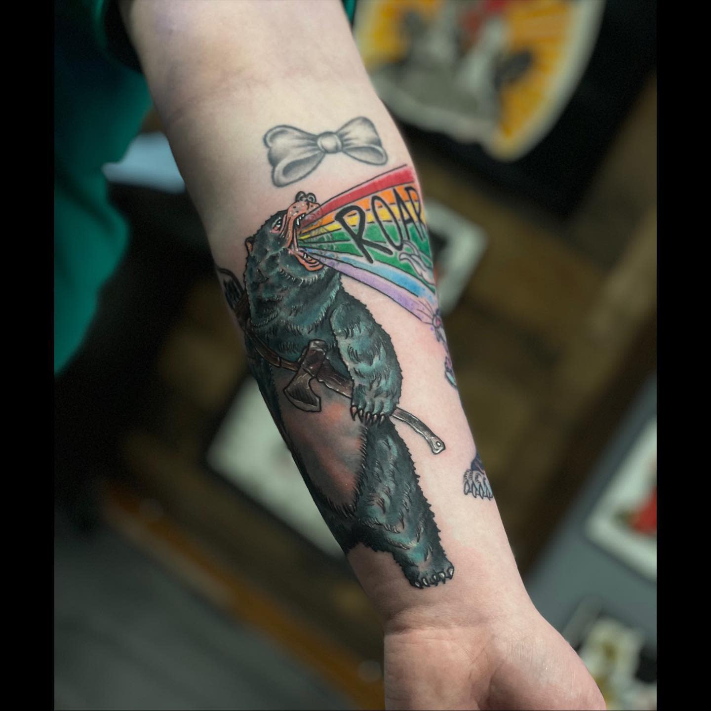 Tattoo of a bear roaring a rainbow with the word "ROAR" on a forearm