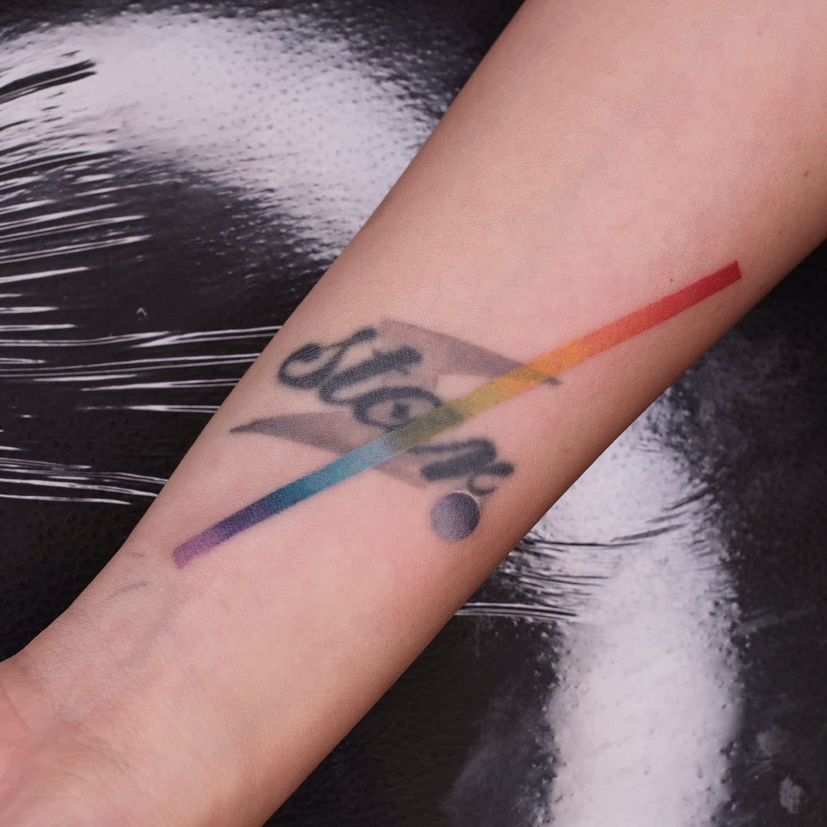 Forearm tattoo of the word "stop" with a diagonal rainbow stripe crossing through it