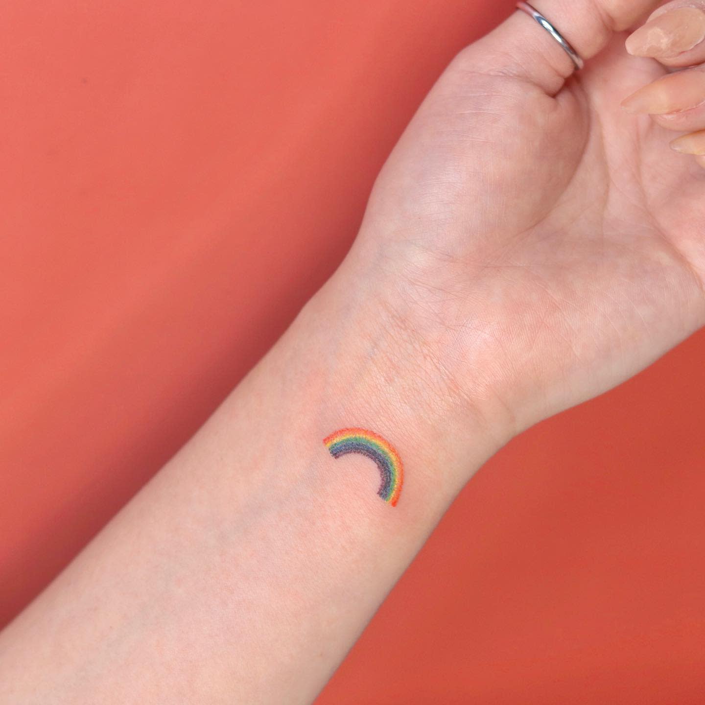 A small rainbow tattoo on an inner wrist against a light orange backdrop