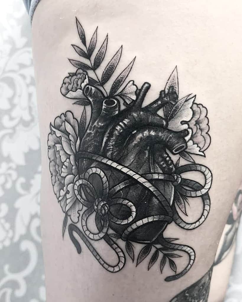 Tattoo of an anatomical black heart adorned with flowers, leaves and ribbon made of rope, featuring detailed shading and linework