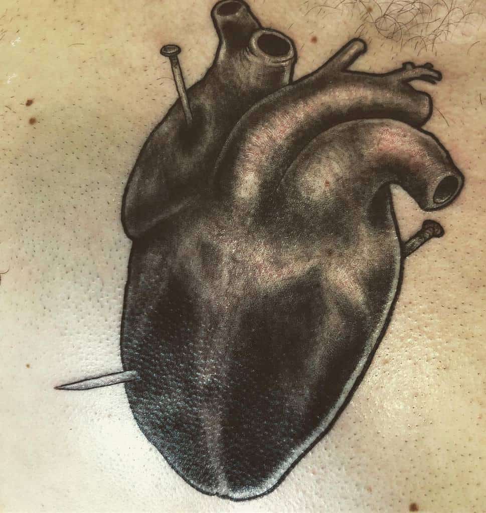 Realistic black and grey tattoo of a human heart with nails piercing it, symbolizing pain or betrayal