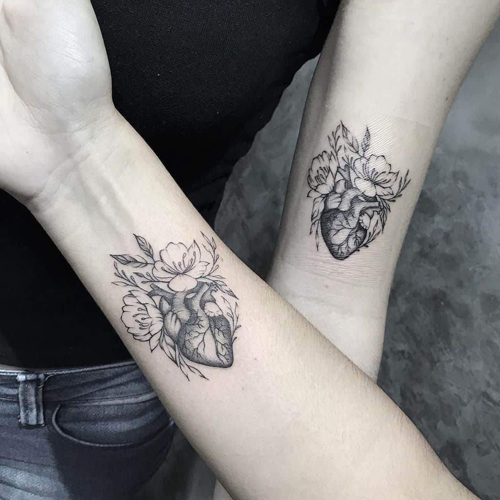 Matching black tattoos on two forearms, each showcasing an anatomical heart surrounded by floral patterns, rendered in fine black line work