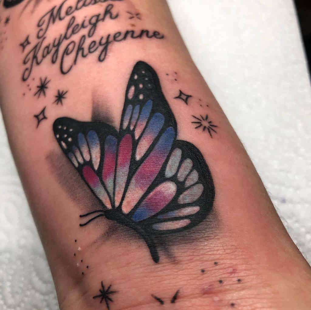 Tattoo of a black butterfly with blue and pink wings surrounded by stars and text on the wrist