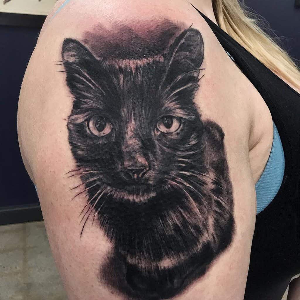 A realistic black cat tattoo on the upper arm, with detailed shading and lifelike eyes capturing the cat's expression
