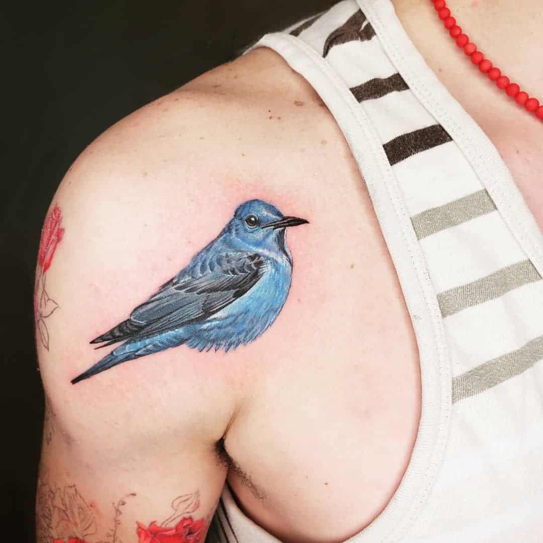 A colorful tattoo of a blue bird on a shoulder wearing a white top with stripes and a red bead necklace