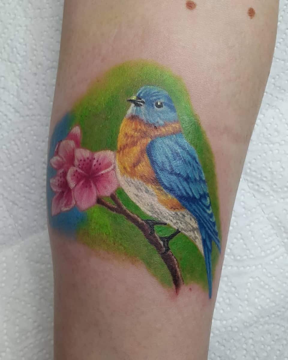 Tattoo of a blue and orange bird on a branch with pink flowers against a green background on an arm