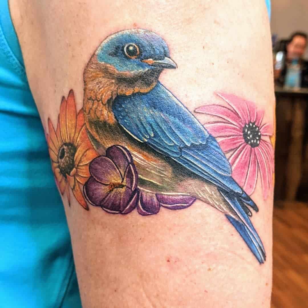 Colorful tattoo of a bluebird perched on a branch with pink and yellow flowers on an upper arm