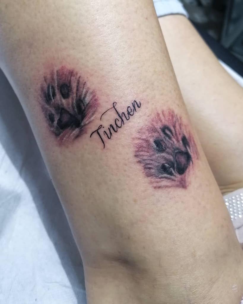 Tattoo of two cat paw prints with the name "Tinchen" in cursive between them on a person's leg