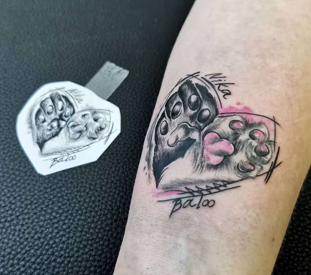 A tattoo featuring a heart-shaped cat paw print with the names "Nika" and "Baloo" on an arm next to a stencil