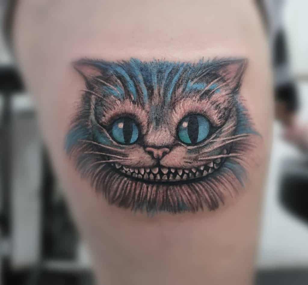 A vibrant tattoo of the Cheshire Cat with blue and grey fur, featuring its iconic wide grin and bright eyes