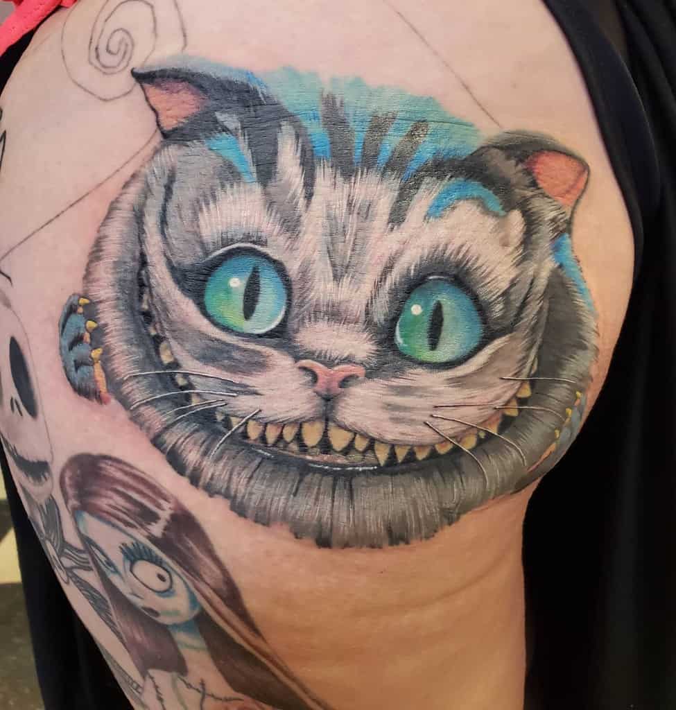 A colorful tattoo of the Cheshire Cat with piercing green eyes, a wide grin, and blue accents on its fur