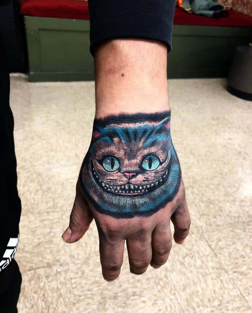 A vivid Cheshire Cat tattoo on the hand, showcasing its wide grin and blue-green eyes with bold colors
