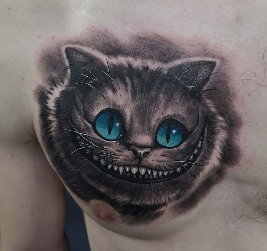 A black and grey tattoo of the Cheshire Cat with vibrant blue eyes, wide grin, and detailed shading on the chest
