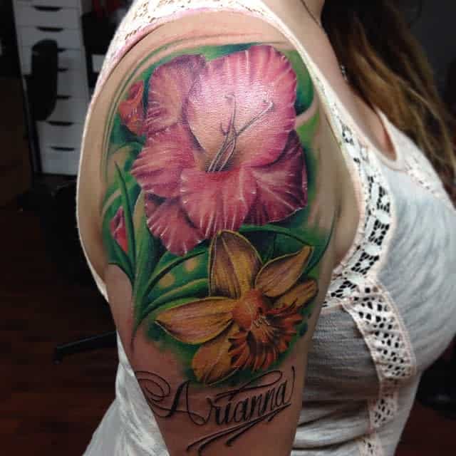 A shoulder tattoo of pink and yellow flowers, including a delicate gladiolus, with the name "Arianna" elegantly written underneath