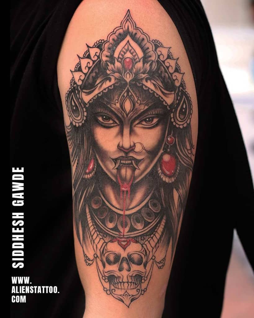 Black and gray tattoo of Kali with a third eye, sticking out her tongue, wearing a detailed headdress, adorned with jewels and skulls