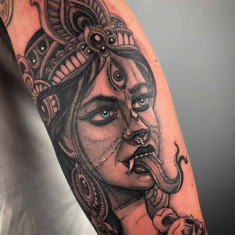 Black and gray tattoo of Kali with a third eye, sticking out her tongue, adorned with intricate jewelry