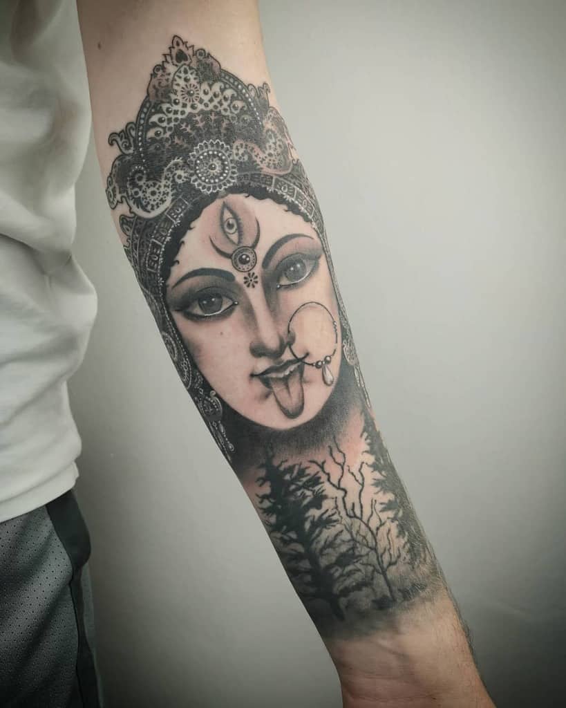 Black and gray tattoo of Kali with a third eye, sticking out her tongue, intricate headdress, and a forest landscape beneath