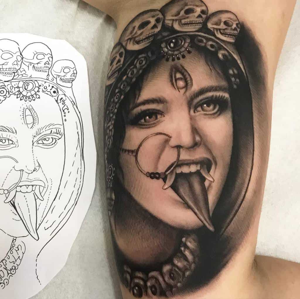 Black and gray tattoo of Kali with a third eye, sticking out her tongue, adorned with skulls and intricate details
