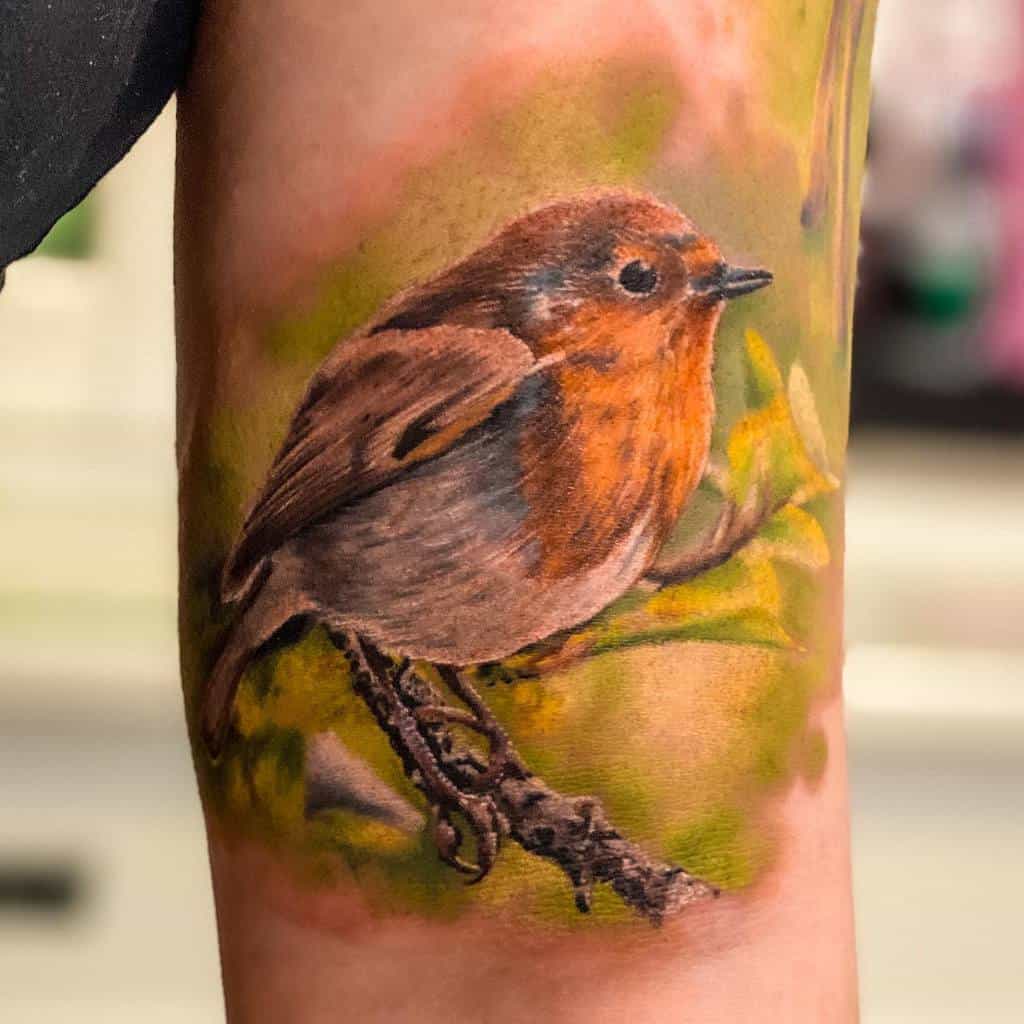 A colorful tattoo of a robin perched on a branch with realistic leaves, blurred green and brown background