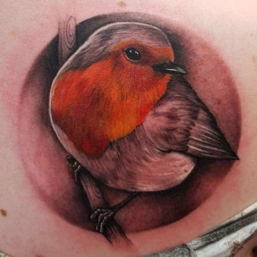 A realistic tattoo of a robin with red and orange breast, perched on a branch, surrounded by a circular shaded background