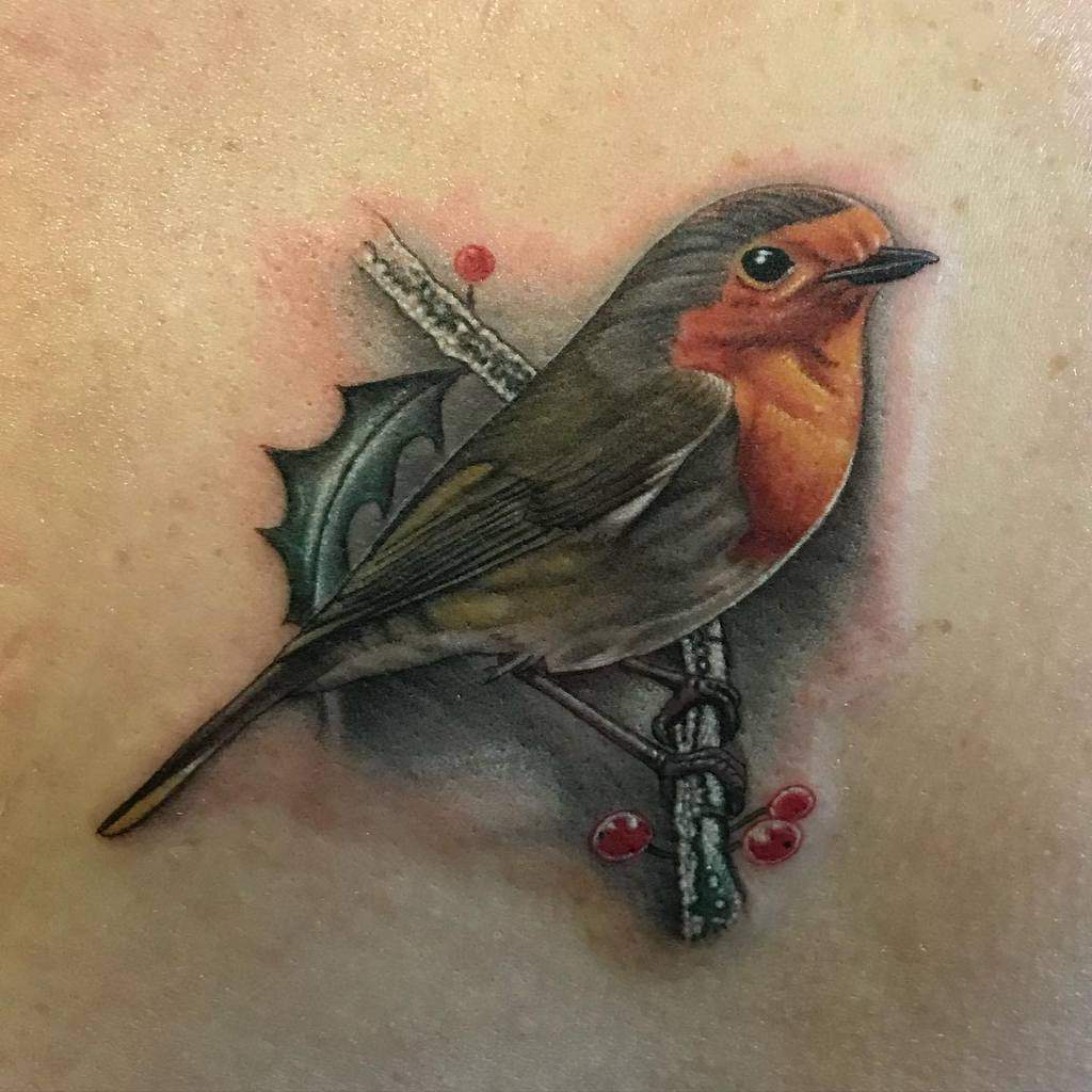 Tattoo of a realistic robin perched on a branch with holly leaves and red berries behind it
