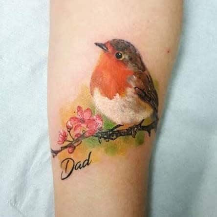 Tattoo of a robin on a branch with pink flowers, the word "Dad" is written below, with green and yellow background
