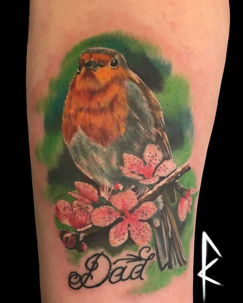 A realistic tattoo of a robin perched on a branch with pink flowers and berries, with the word "Dad" written below, on a green background