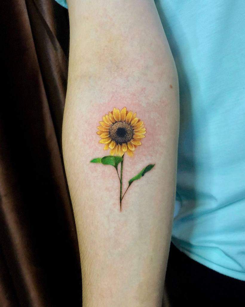 A tattoo of a sunflower on a person's arm, featuring a brown center and yellow petals with a green stem and leaves