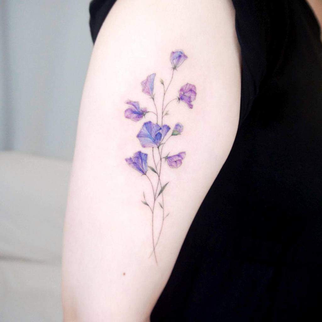 A realistic tattoo of sweet pea flowers in soft purple tones, gracefully placed on the upper arm