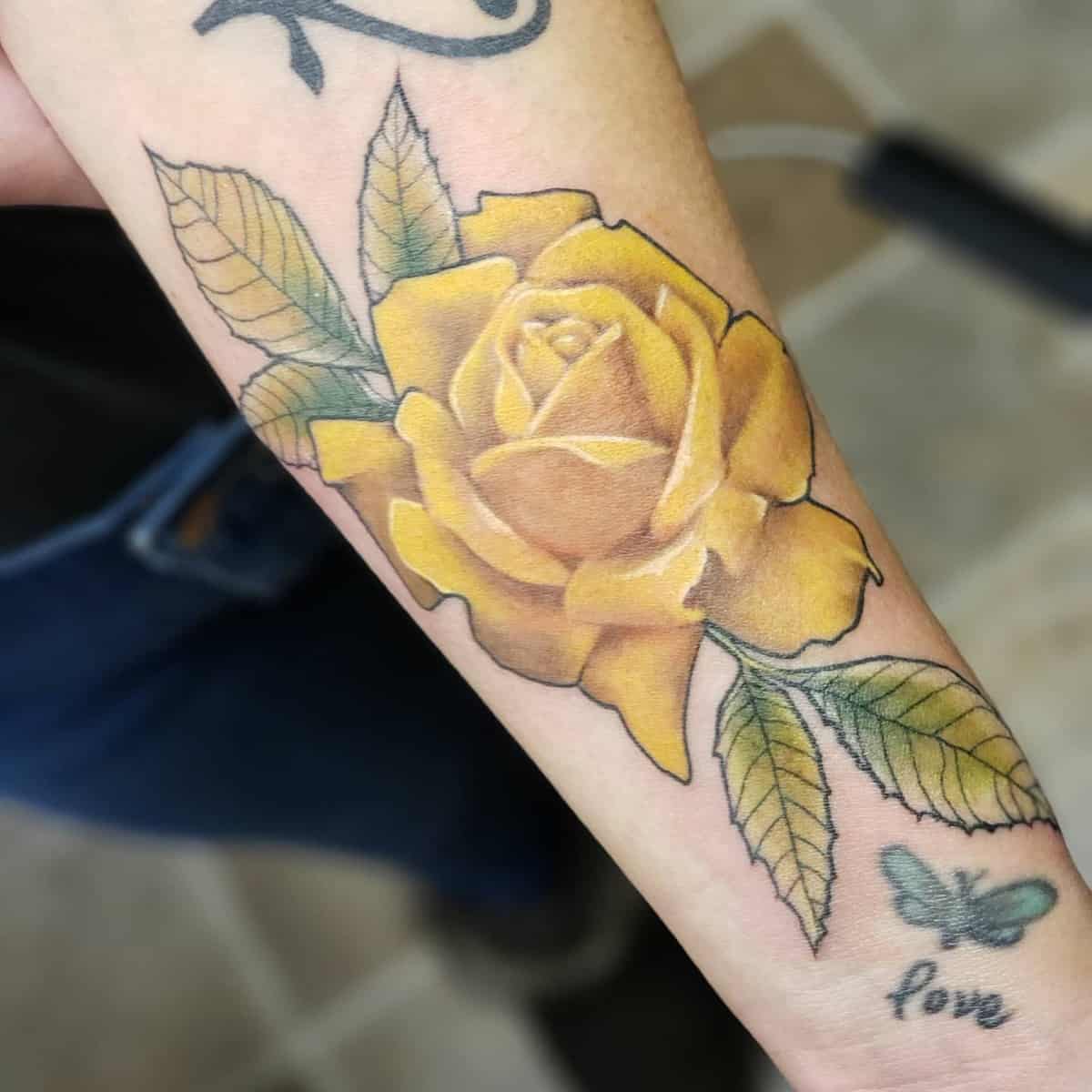 A tattoo of a yellow rose with green leaves on a forearm with a small tattoo of the word "love" and a butterfly nearby