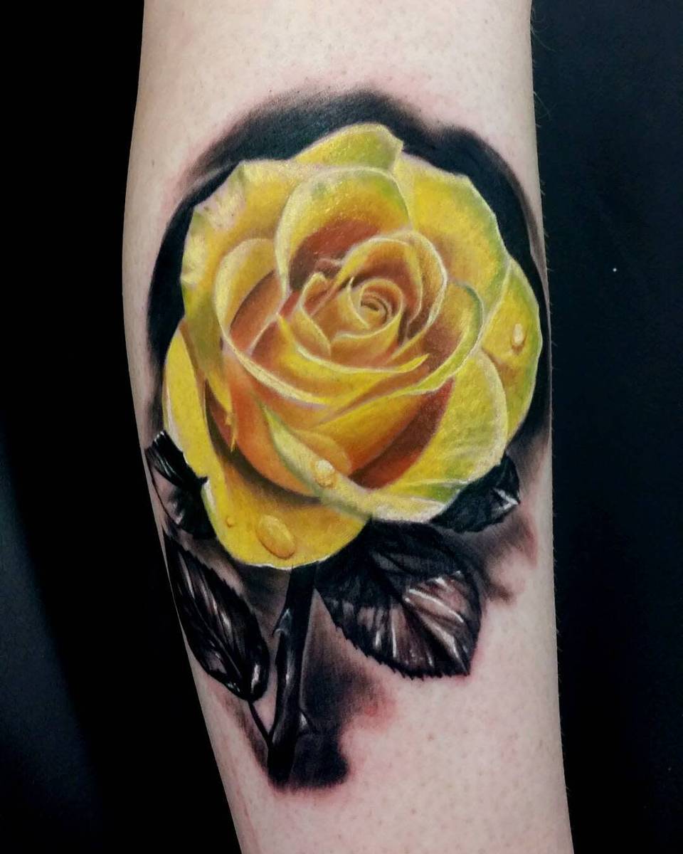 A realistic yellow rose tattoo with water droplets on a forearm