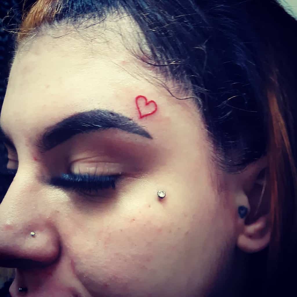 Small red heart outline tattoo above the eyebrow