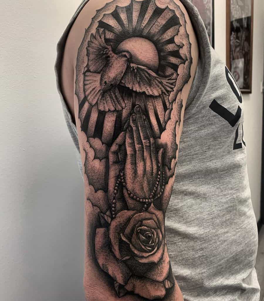 Religious-themed tattoo featuring praying hands holding rosary beads, a rose, a dove, and rays of light shining from behind clouds