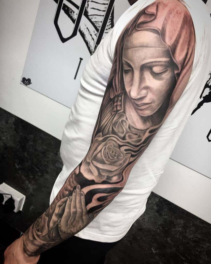 A religious-themed full sleeve tattoo in black and grey featuring a detailed portrait of the Virgin Mary, roses, and praying hands holding rosary beads, extending from the shoulder to the wrist