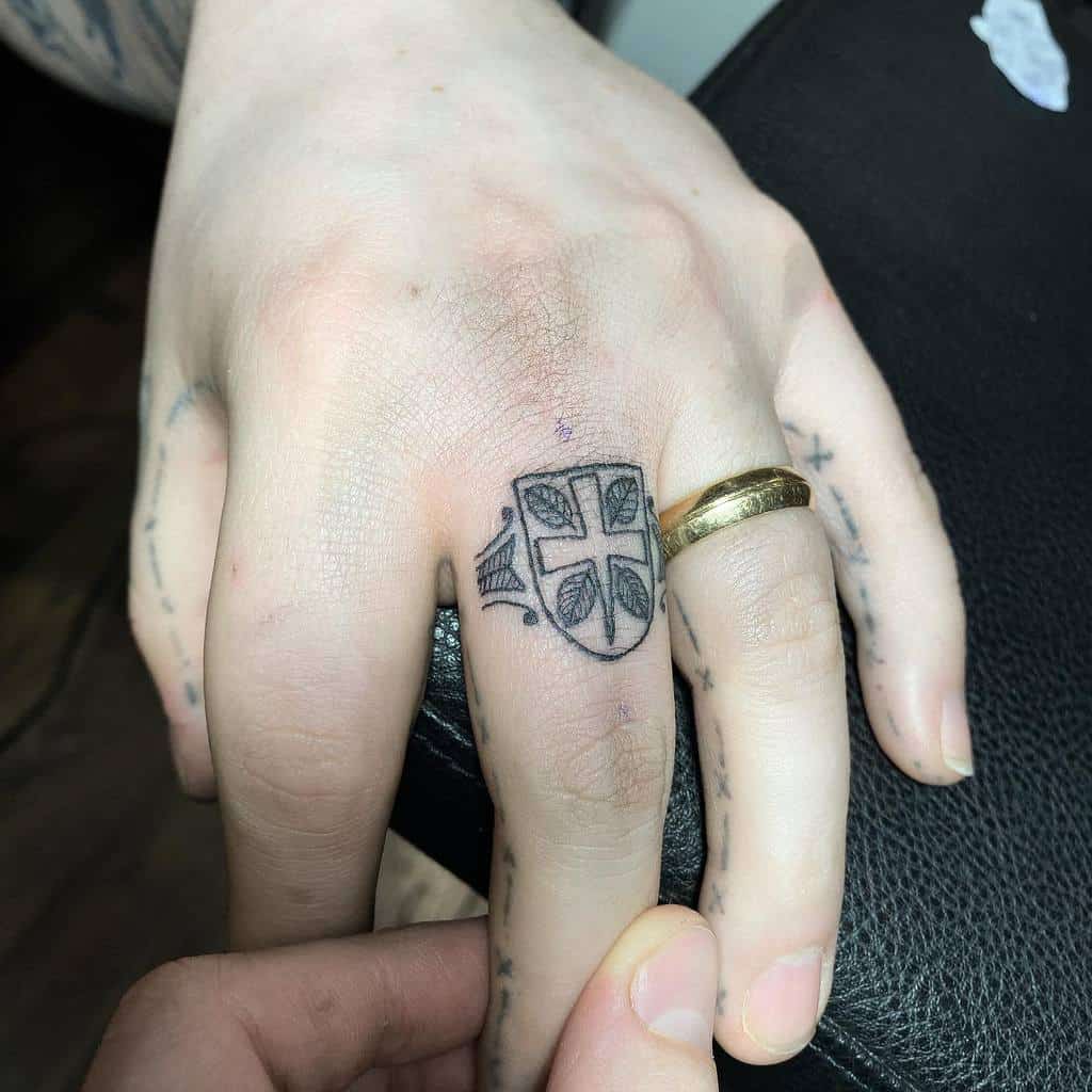 A black ink ring tattoo of a shield with a cross and leaf-like patterns