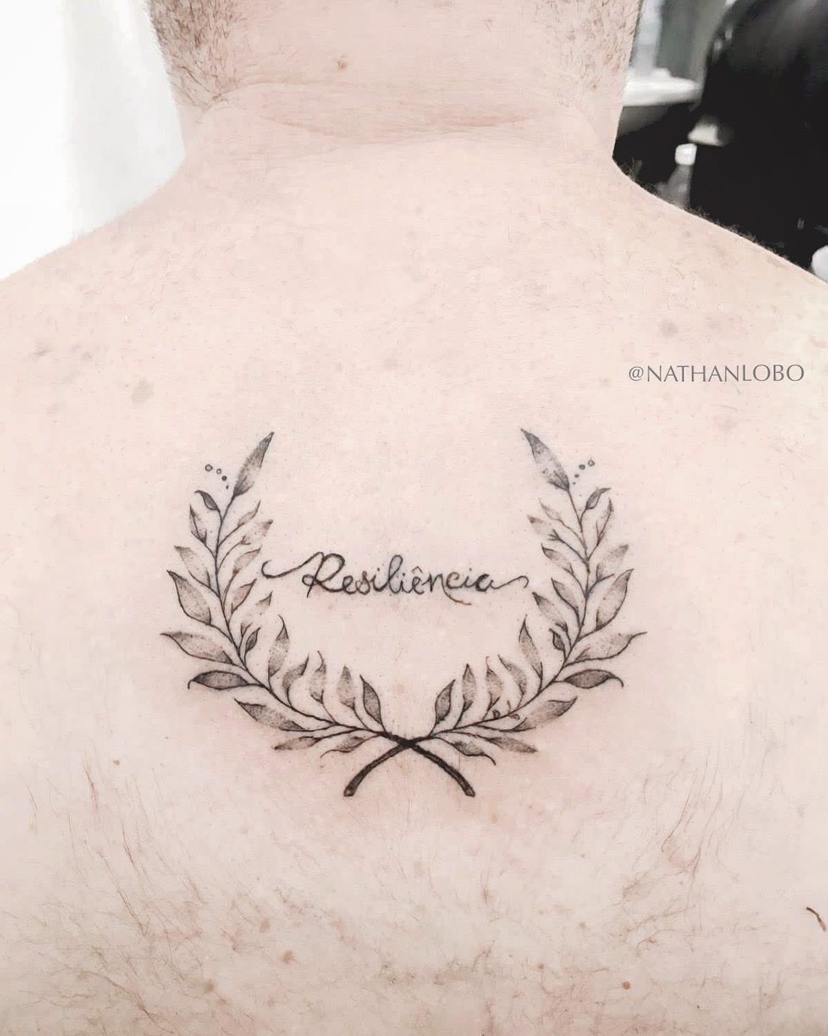Tattoo of "Resiliência" with laurel wreath design on upper back