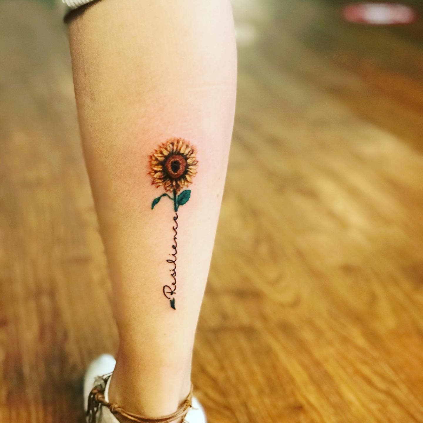 A sunflower tattoo on a leg with the word "Resilience" as the stem