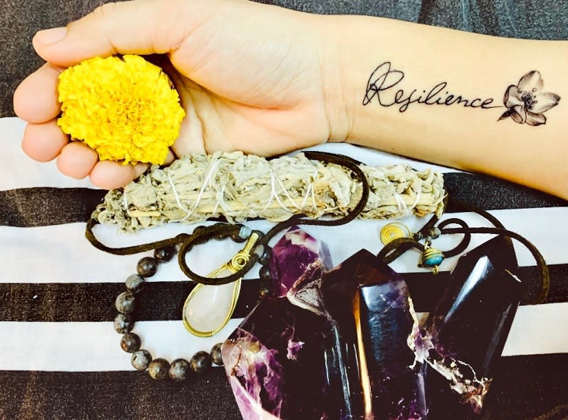 A hand holding a marigold flower and sage bundle with "Resilience" tattoo and crystals
