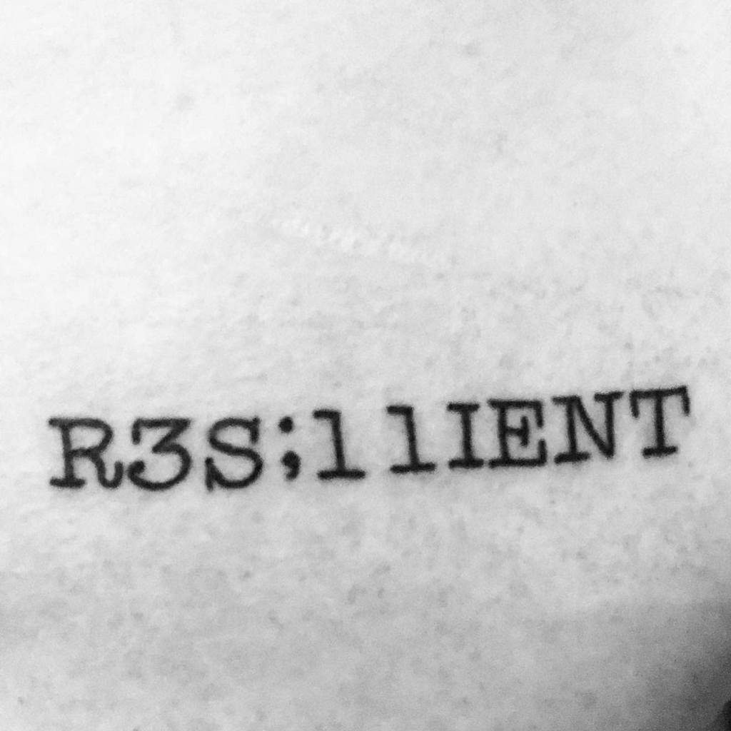 This tattoo features the word "Resilient" with some stylized elements, using numbers and punctuation marks creatively in place of certain letters, adding a unique touch to the concept of strength and perseverance.