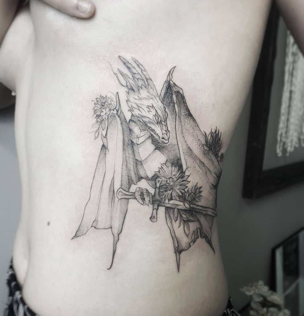 Black and grey tattoo of a dragon holding a sword, with flowers, on a woman's side