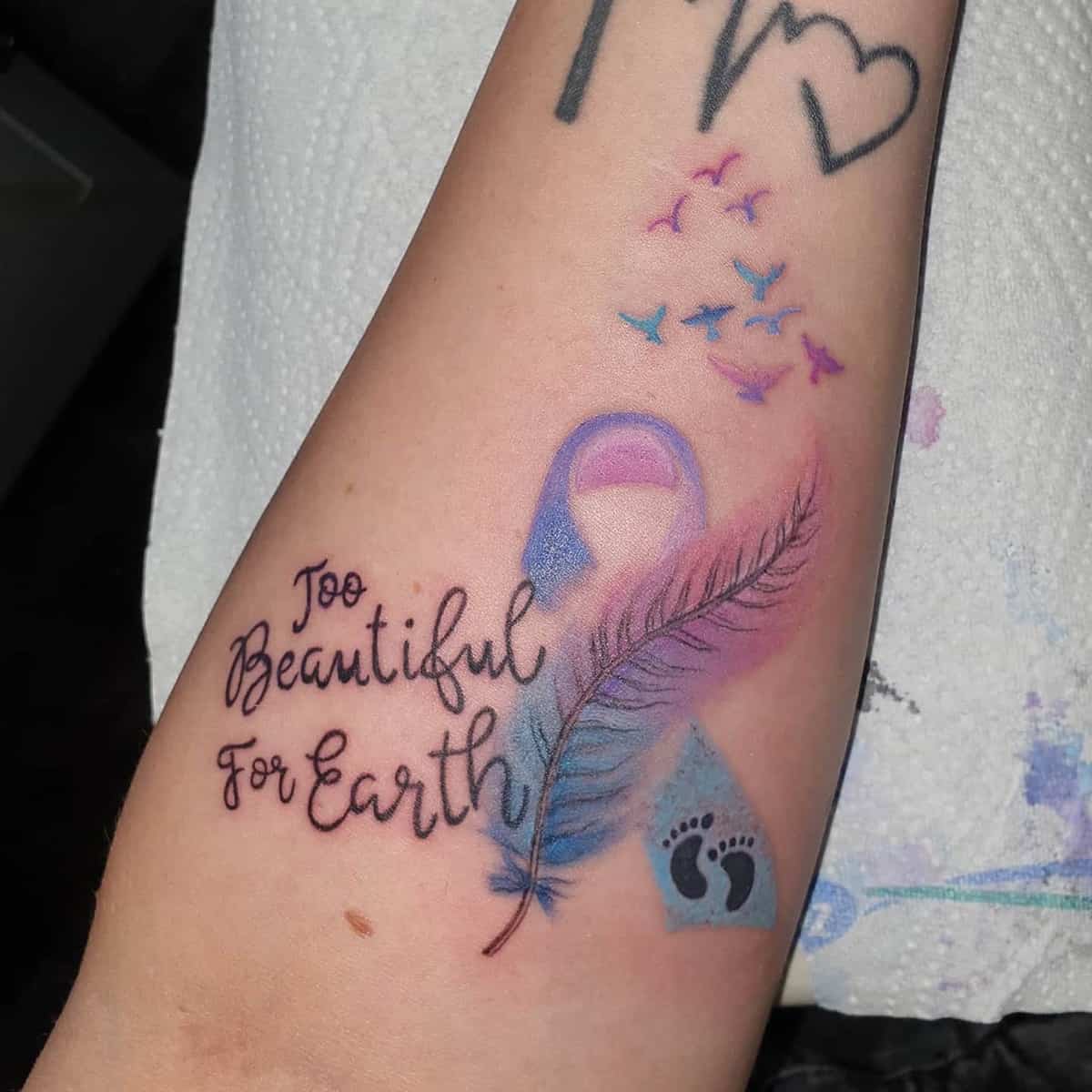 A tattoo featuring a pink and blue ribbon, a feather, birds, footprints, and the words "Too Beautiful For Earth"