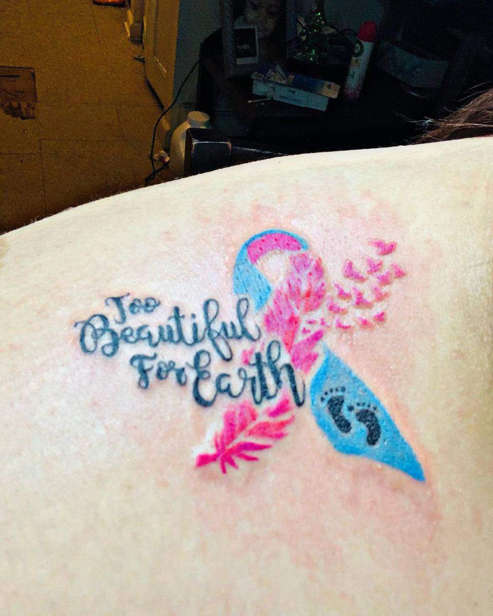 Tattoo of a pink and blue ribbon with wings, feather, baby footprints, and "Too Beautiful for Earth" text on skin
