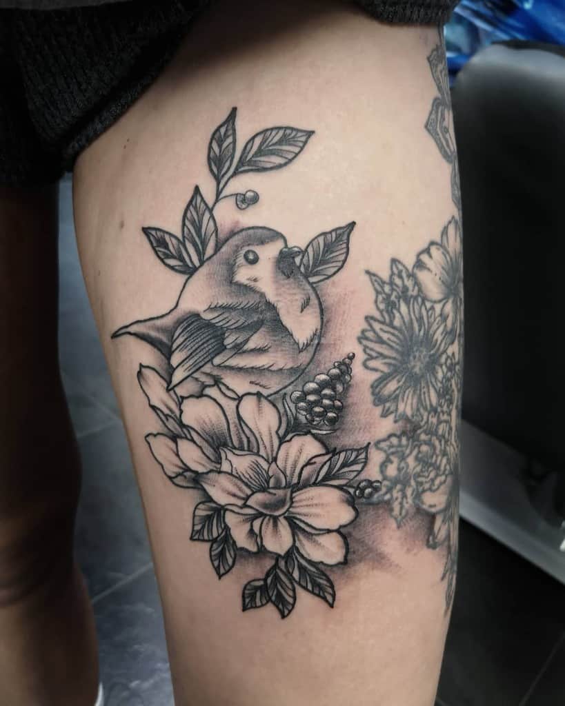 Black and grey tattoo of a robin perched among detailed flowers, leaves, and berries, featuring fine linework and intricate shading