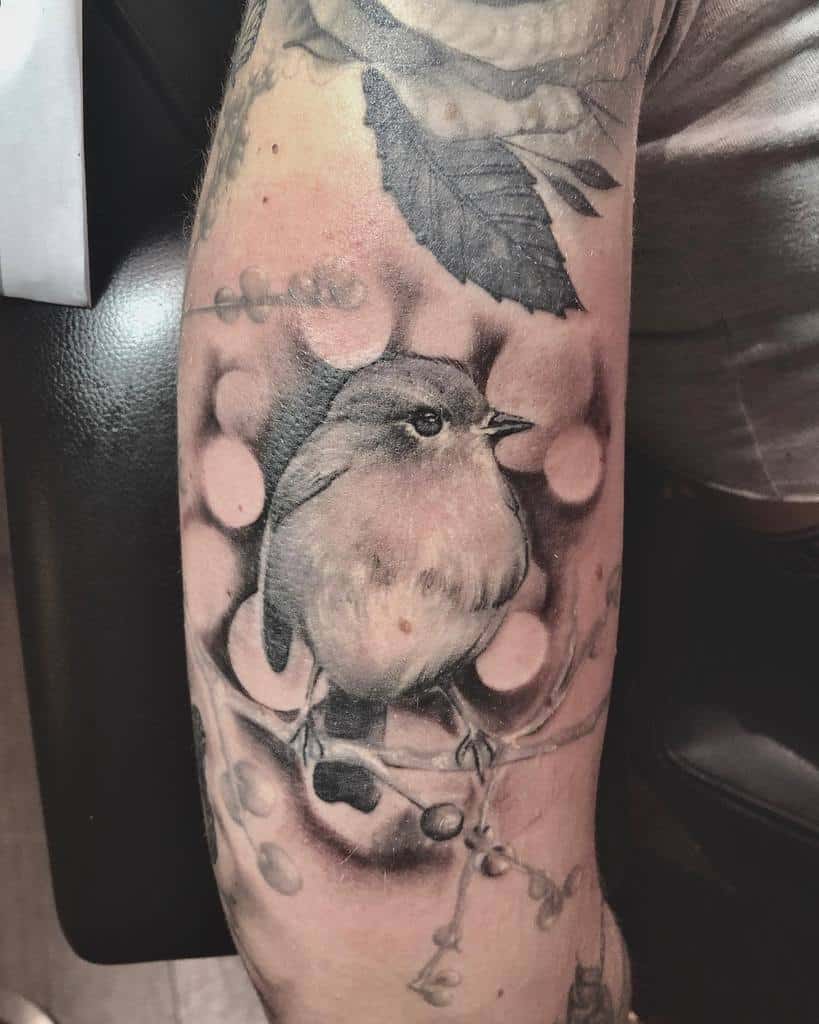 Realistic black and grey tattoo of a robin perched on a branch with berries, with detailed shading, soft highlights, and a blurred bokeh-style background
