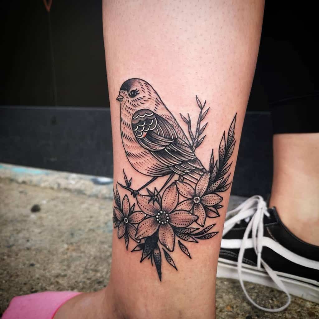 Black and grey tattoo of a robin perched on a branch, surrounded by detailed flowers and leaves, featuring fine linework and intricate shading
