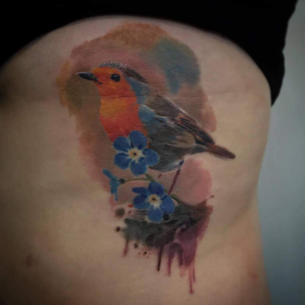 Colorful tattoo of a robin with red and orange plumage perched on branch with two blue flowers, set against a brownish watercolor background