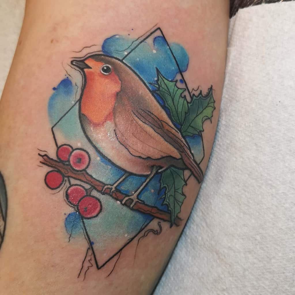 Tattoo of a robin perched on a branch with holly leaves and berries, against a diamond shape blue watercolor background