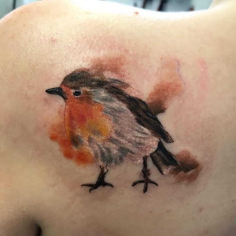 Watercolor-style tattoo of a robin with soft orange hues on its chest, surrounded by splashes of warm color, creating a fluid and artistic design