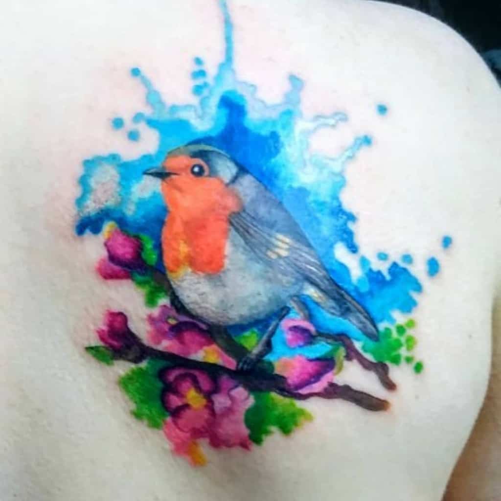 Tattoo of a colorful robin perched on a branch with pink flowers and leaves, set against a blue splash background