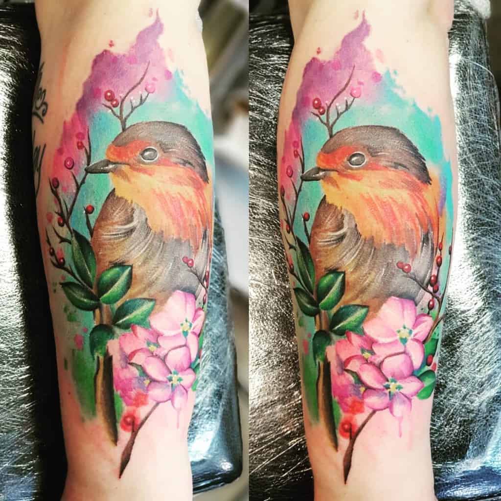 Two images of a colorful tattoo of a small robin with vibrant orange and brown plumage, surrounded by pink flowers, red berries and green leave, with blue and pink watercolor background